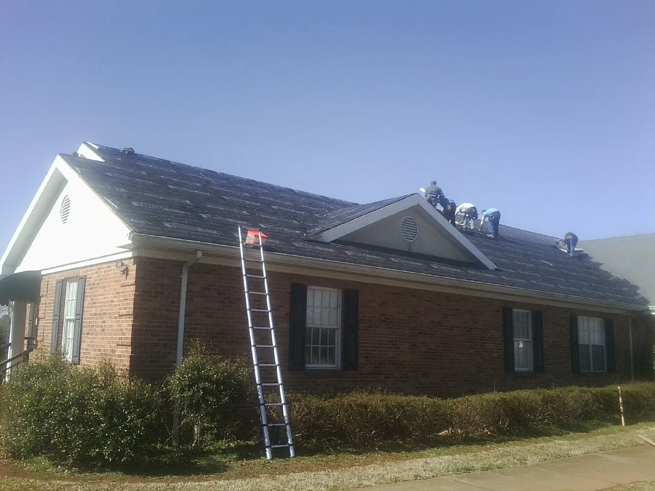 Crew tearing shingles off a brick ranch roof with a ladder set — Metal Roof Repair work by our crew in Stoneville