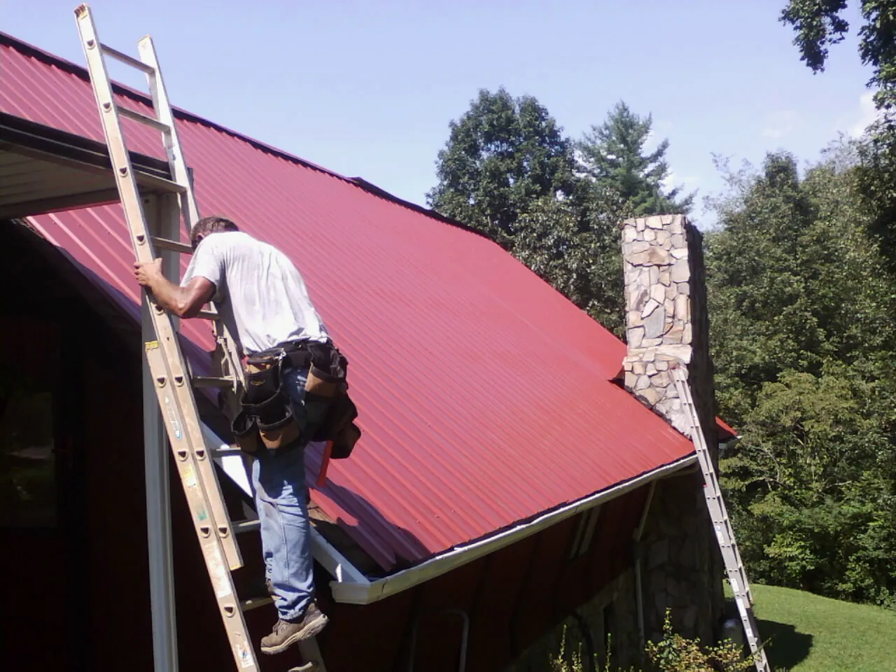 Roofer on a ladder beside a red metal roof and stone chimney — Soffit & Fascia Repair by Roofing Brothers in Stoneville
